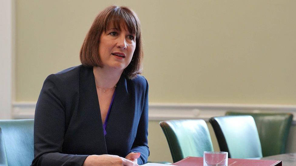 Rachel Reeves sits at a desk with empty chairs beside her. She's leaning forward and appears to be listening and is holding a piece of A4 paper