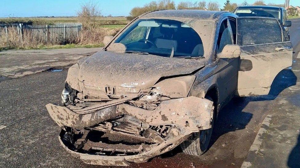 A mud splattered vehicle which has been damaged and crushed at the front underneath the bonnet. The vehicle is parked by the side of a road with a field in the background.