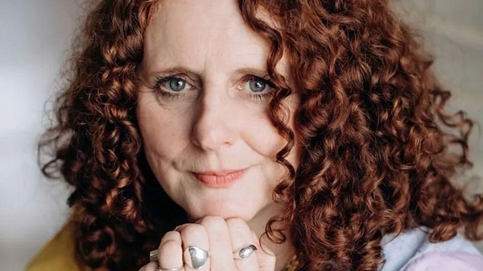Maggie O'Farrell sits with her hand clasped together under her chin. She has red curly hair and blue eyes.