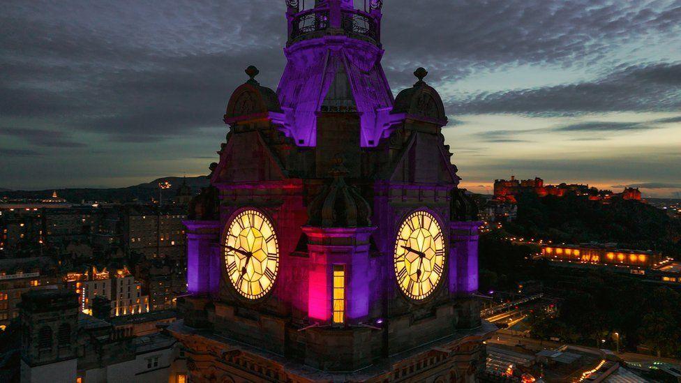 A close up of the Balmoral Hotel clock showing two of its faces. It is lit up in a purple light and looks atmospheric because it is against a dark nights sky. In the background are the lights of the city's streets and Edinburgh Castle.