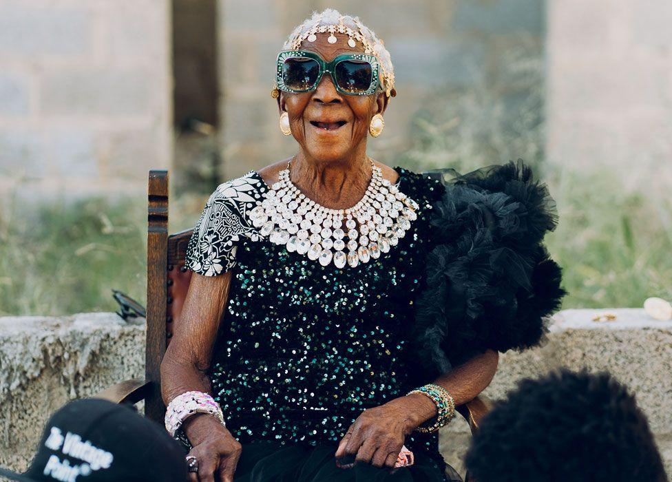 A seated Margret Chola laughs at the camera. She is wearing an Indian-style  head band, big bejewelled green-framed sunglasses, a diamond-like waterfall necklace and a black feather boa over a top covered in sequins