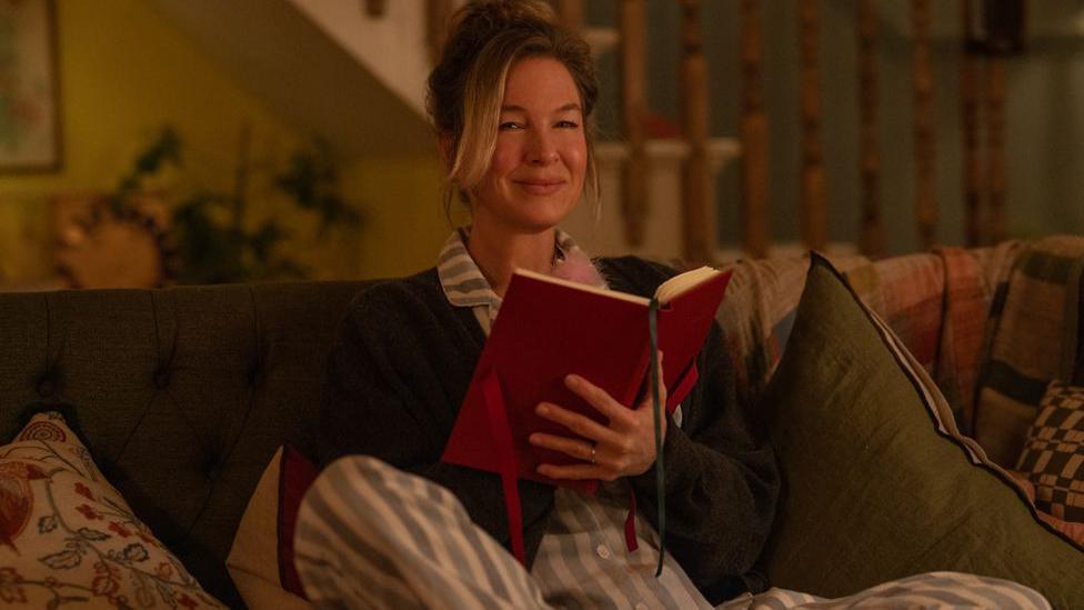 Renee Zellweger as Bridget Jones in the fourth Bridget Jones film. She is dressed in white and blue striped pyjamas with a grey cardigan. She is sat on a sofa holding a red diary and smiling. 