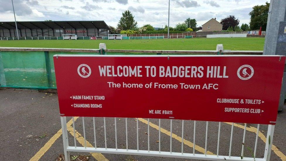 A red sign in the foreground says Welcome to Badgers Hill, the home of Frome Town AFC. Behind the sign is a green football pitch.