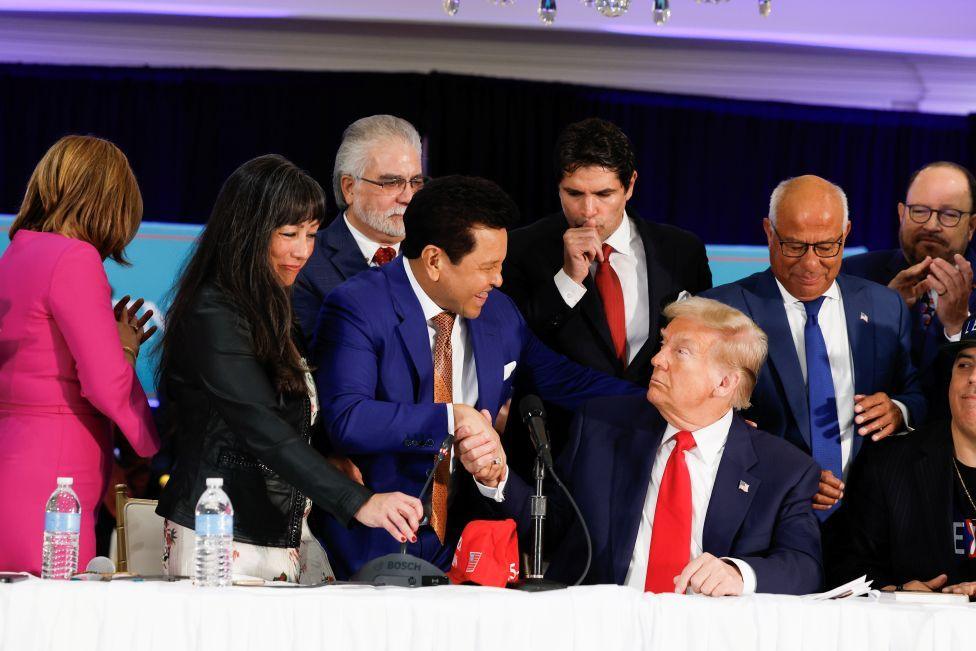 Trump përshëndet njerëzit me shtrëngime duarsh ndërsa ata i afrohen tavolinës së tij. Ai është ulur i veshur me kravatë të kuqe dhe kostum ngjyrë blu benzine dhe ka turma njerëzish përreth tij.