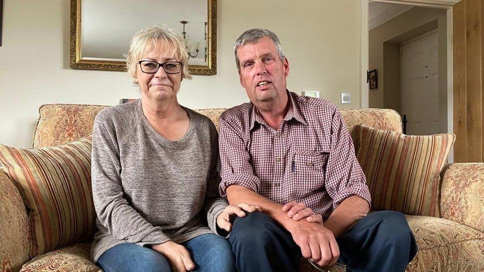 Amanda and Martin Beal are sitting on a brown sofa in their living room. She is wearing jeans and a brown jumper and has her hand on his knee. He is wearing a shirt and jeans.