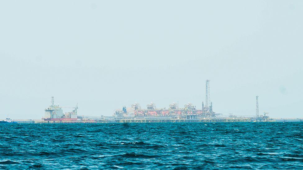 A gas platform out at sea can be seen on the horizon - the ocean can be seen in front of it and above a large view of the sky.