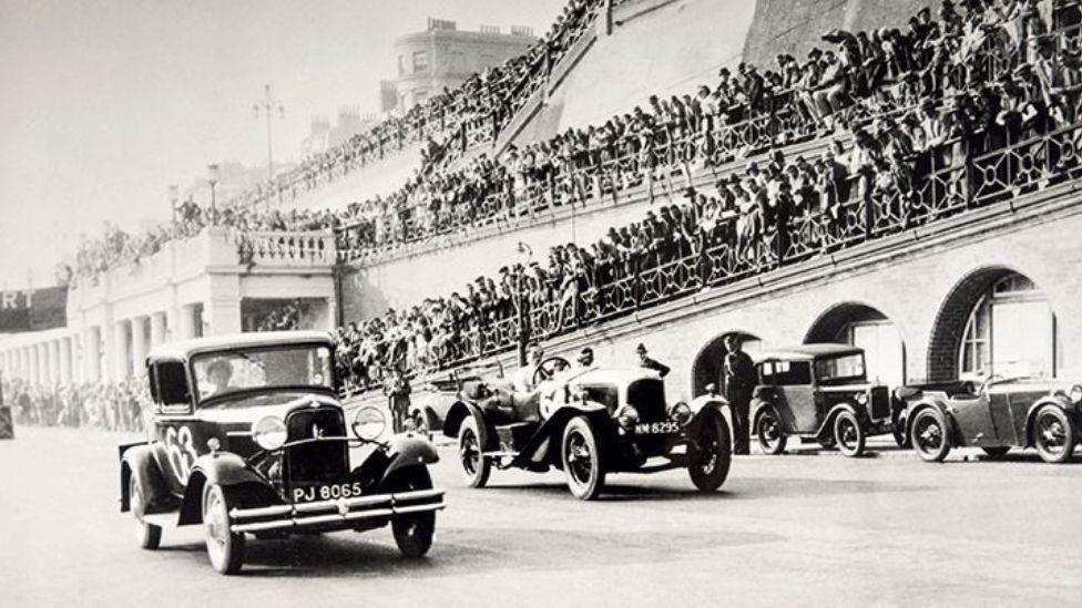 An old black and white photograph of two cars racing on a road, with hundreds of spectators looking on. 