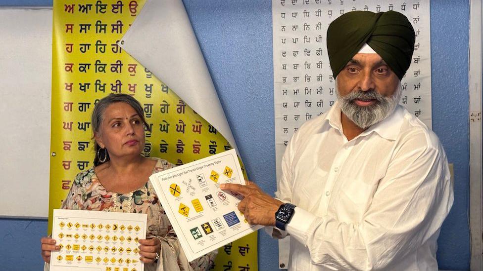An older man wearing a black turban and a white button-down shirt points to a sheet of paper with road symbols on it. Next to him is an older woman in a floral dress, holding a similar sheet with different symbols. They stand in front of two posters with Punjabi lettering