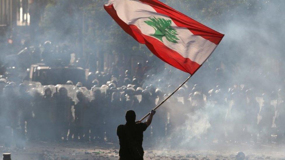 A man holds a Lebanese flag near riot police at a protest on 8 August 2020