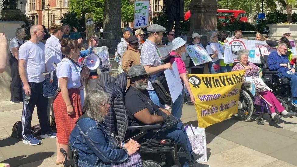 A crowd of around 30 protesters of varying ages stand in a protest. They are all looking to the left and two are holding a yellow banner reading "WinVisible". Two people in the foreground are wheelchair users. Buildings and a red London bus can be seen in the background.