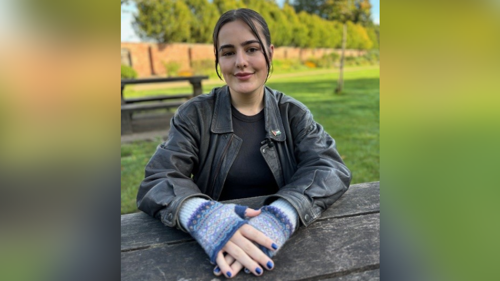 Manon Chiswell is looking at the camera. She is sat on a picnic table with her hand on the table itself. She has blue nail varnish and blue fingerless gloves. She is wearing a black leather jacket under a black t-shirt.