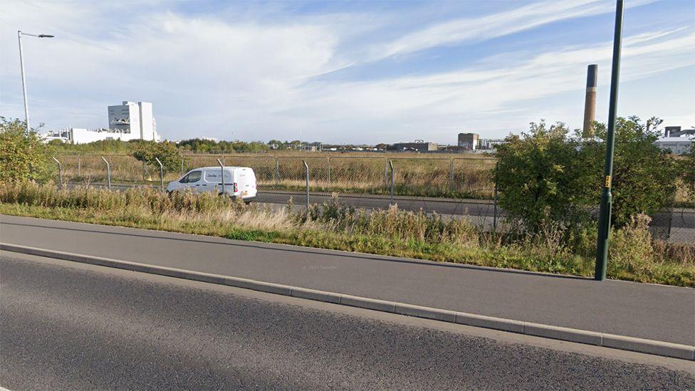 Energy Park Way in Grimsby showing a road with a solitary white van driving along it with chain link fences running alongside it and fields and factories in the background