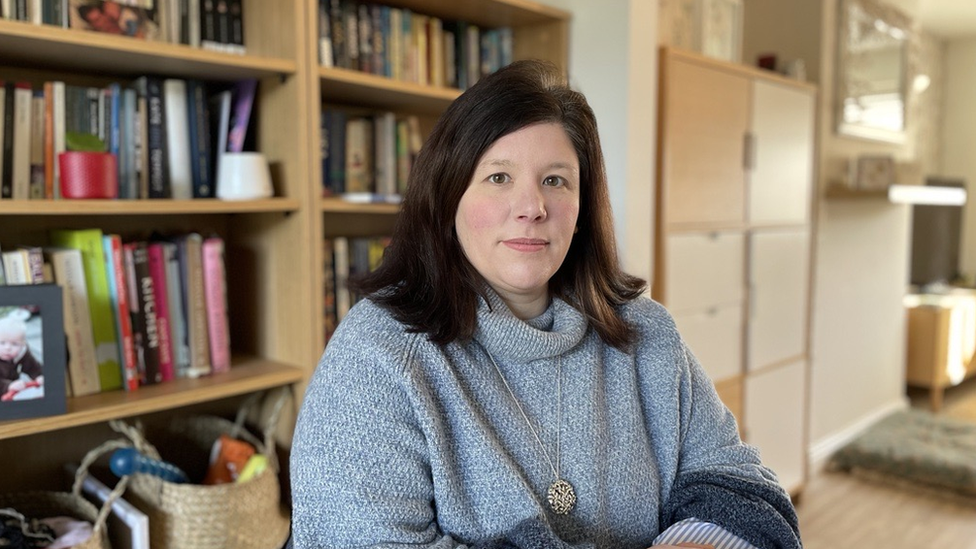 A woman sat in a room with book shelves behind her. She is looking at the camera and wearing a grey jumper.