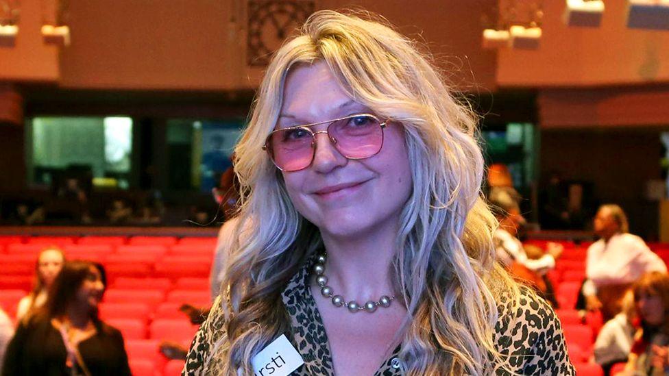 Kirsti wears pink-tinted glasses and a leopard print shirt. She is smiling into the camera while in wht looks like an auditorium with people and red seats in the background