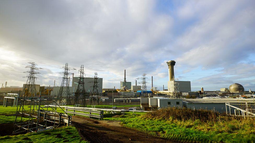 An exterior shot of Sellafield Cumbria the nuclear site where 140 tonnes of plutonium is currently stored The picture shows green grass and scrub in the foreground and a little further back an industrial plant with pylons and silver-grey towers and a large round structure in the background against a mottled sky of clouds with some blue sky and sunshine poking through