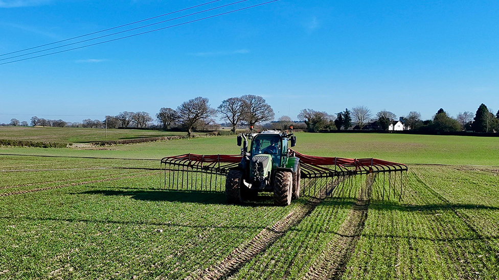 A tractor in a green field, with a red metal attachment spreading out from both sides and a blue sky overhead