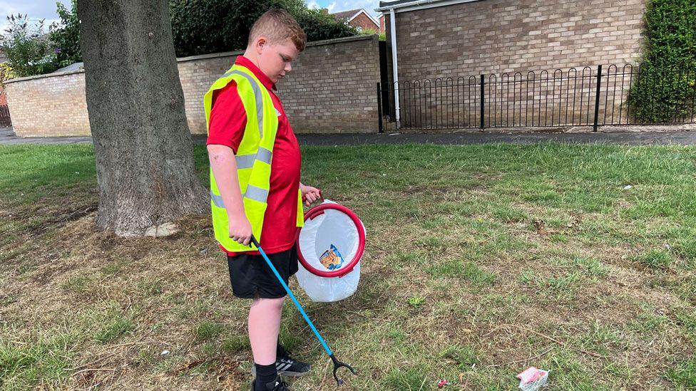 A boy aged 12 standing on grass with a tree behind him and a garden wall. He is looking down at the grass wearing a hi-vis yellow jacket over a red polo-neck and black shorts. In his left hand is a litter picking hoop and plastic bag and in his right is blue-handled litter picker. On the grass is a piece of rubbish.