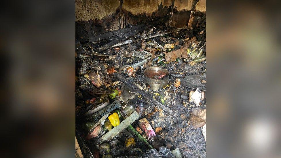 The burnt remnants of a kitchen. There are pots on the floor and some peppers. Everything is broken and burnt and covered in black ash.