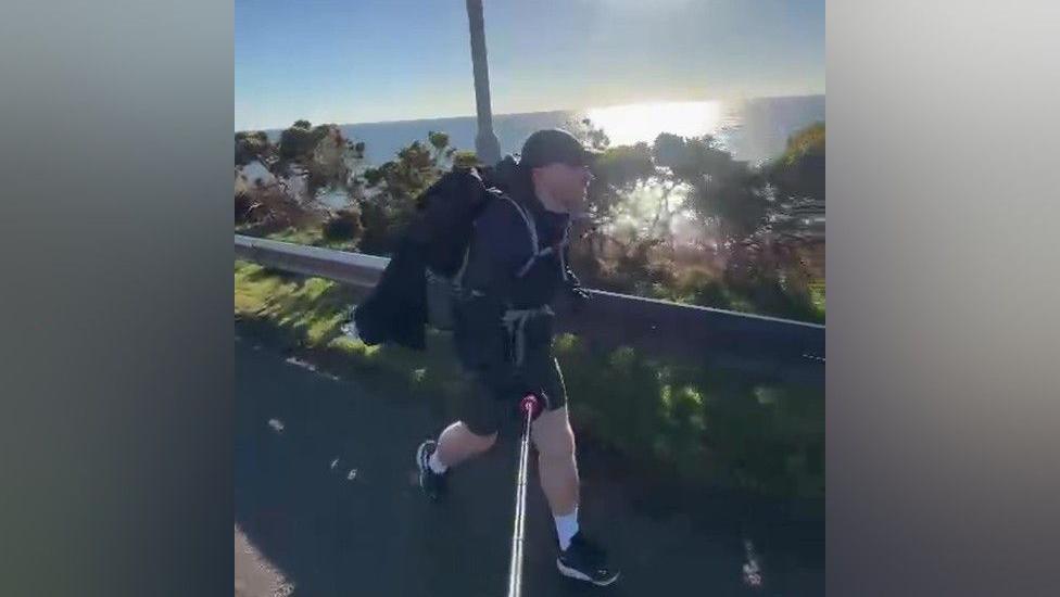 Mr Ward is in a black jacket, shorts and black cap. He is carrying a black backpack. He is running on a road, with grass and bushes behind him. Beyond that is the sea, with the sun reflecting off it.