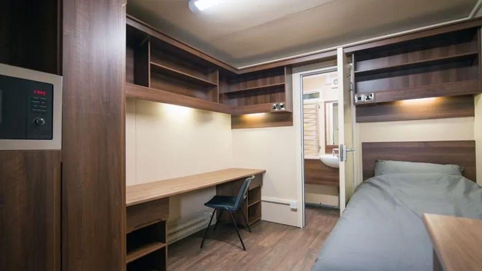 The inside of a pod with dark wood shelving, a desk and chair to one side and a single bed to the other, with a door in the middle going through to a small bathroom