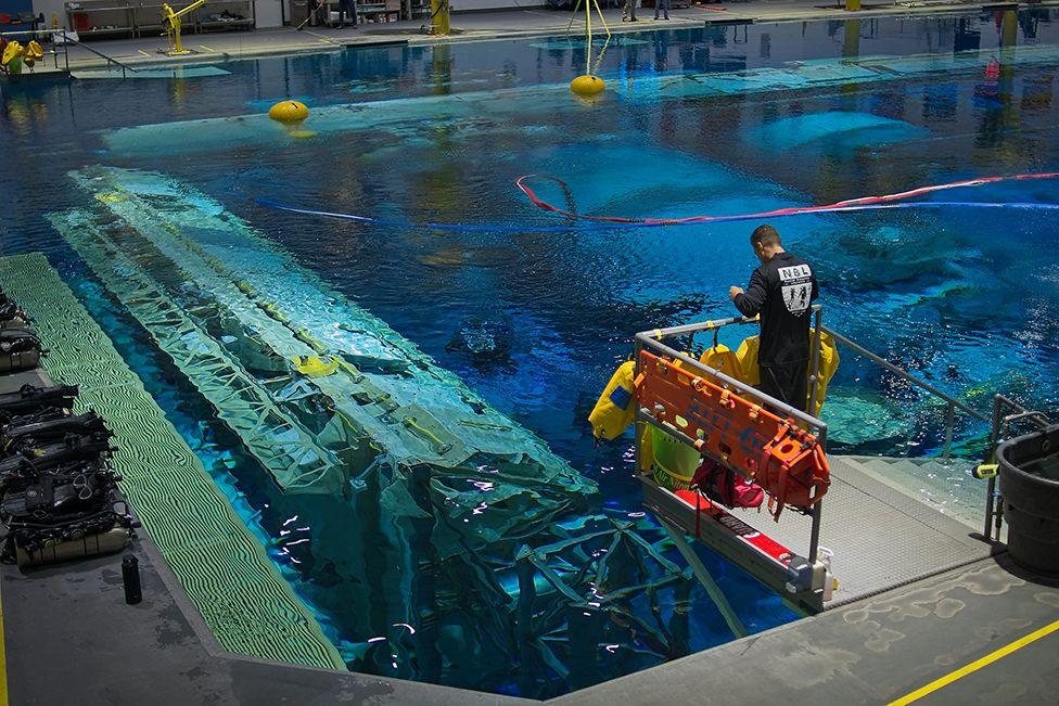 Wide of support team member standing on a platform overlooking the water of the pool. Some of the modules of the space station are clearly visible below the surface. There is a row of scuba equipment to the left of the picture and there are red and blue " umbilical hoses " stretching down to the unseen astronauts beneath the water's surface.