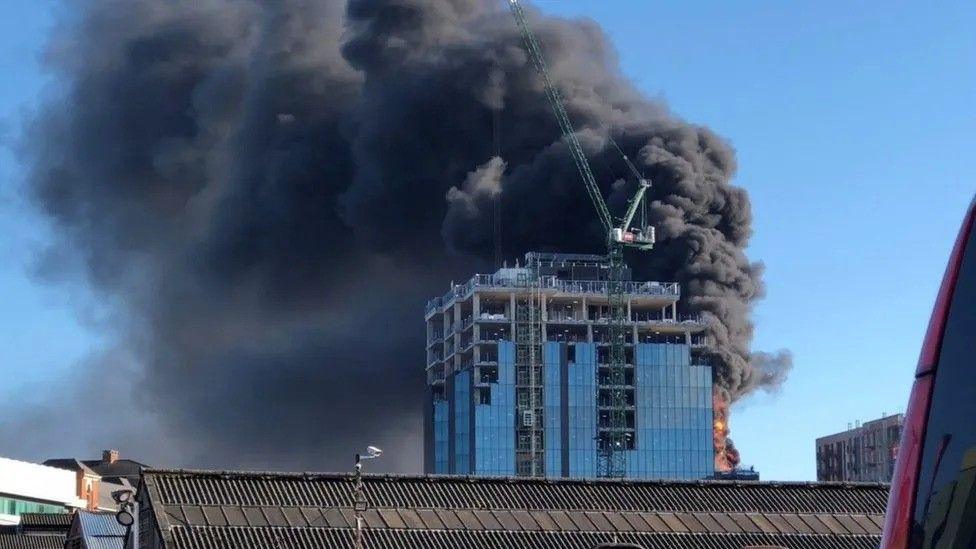 Large plumes of smoke bellow out from a high-rise building which is mid-construction. The crane is in the foreground with the cage attached.