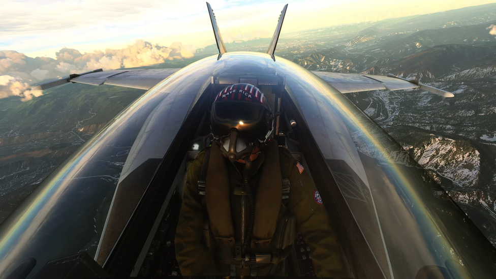 A pilot with a helmet reading "Maverick" sits in the cockpit of a fighter plane, the world behind him