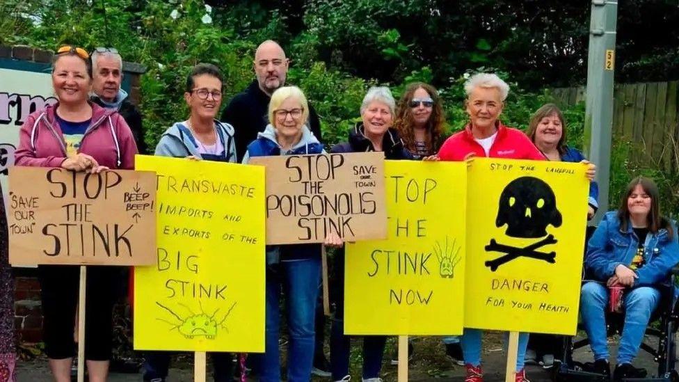 A group of protesters, including MP Lorraine Beavers holding bright yellow placards saying "stop the stink". There are ten protestors in all, with nine standing behind placards and one lady seated in a wheelchair.