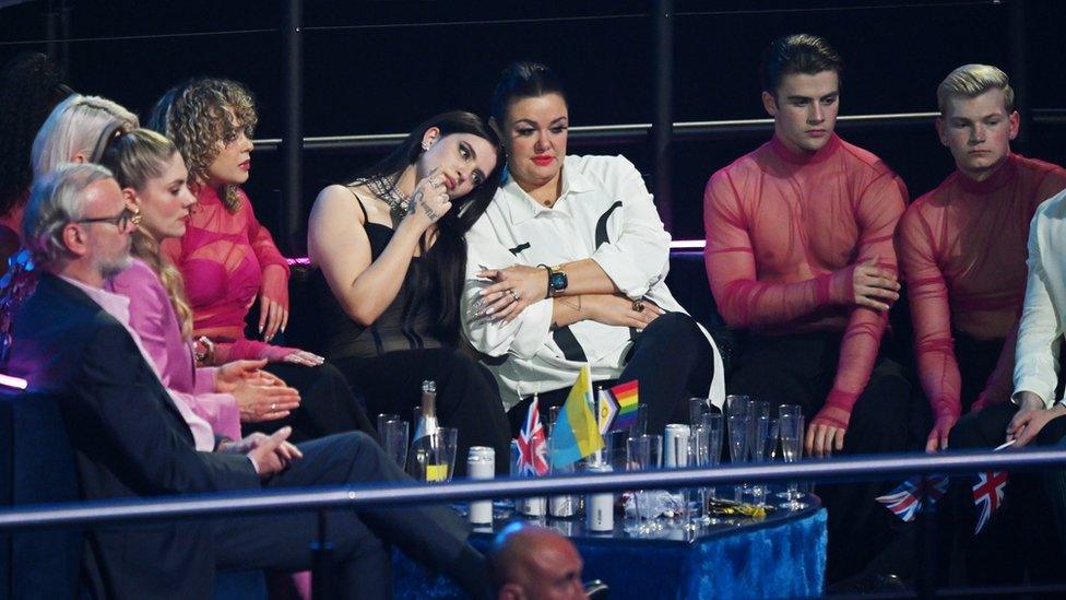 Mae Muller reacts to the UK's low score in the Eurovision green room. She's wearing her stage outfit, and looks glum as she rests her head on another contestant's shoulder. She's surrounded by other entrants in their performance clothes, and the mood is tense as they all watch a monitor as scores are announced.