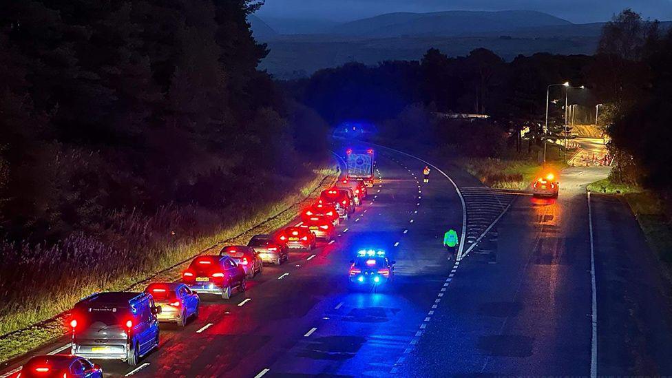 Vehicles on the M6 northbound carriageway, near Tebay west services being diverted near junction 38 after the fatal crash. It is dark and the car lights are bright red and blue. 