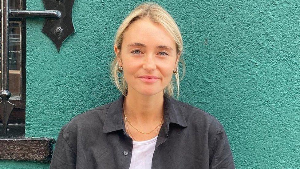 Billie Croucher, who is blonde and wearing a black shirt, standing against a green wall