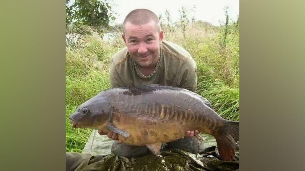 Terry Ricketts, pictures holding a large fish he has just caught. His hair is cut very short and he wears khaki-coloured clothes to blend in with the river bank or lakeside which is green and overgrown behind him. 