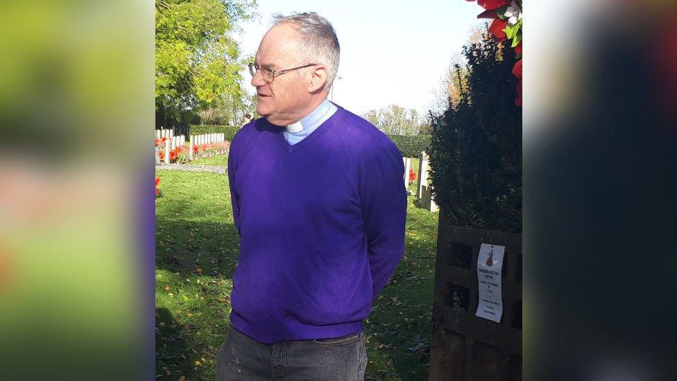 A man wearing a purple jumper, a priest's dog collar and black jeans standing in front of a graveyard and looking away from the camera. He has short grey hair and is wearing glasses.