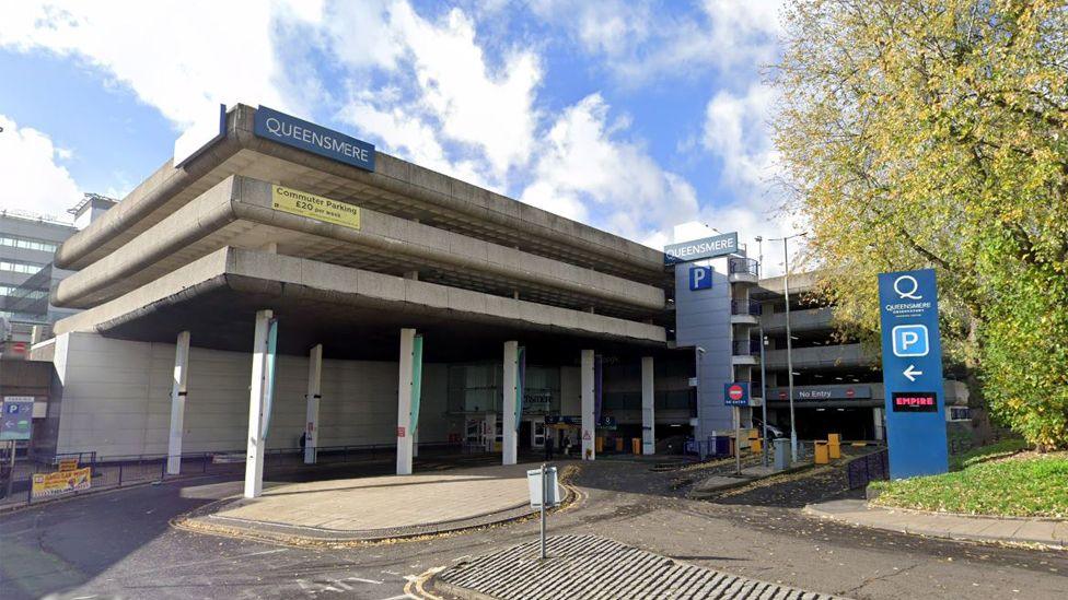 the concrete exterior of Queensmere shopping centre multi storey car park. You can see at least four storeys. There are blue signs with Queensmere in white writing. There are several blue P parking signs and a large tree to the right of the car park.