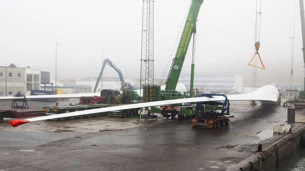 A large wind turbine blade is transported across tarmac by an industrial vehicle. Cranes and industrial buildings are visible in the background