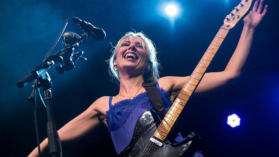 Member of Wolf Alice holding a guitar with her arms outstreched smiling, with a microphone in front of her 