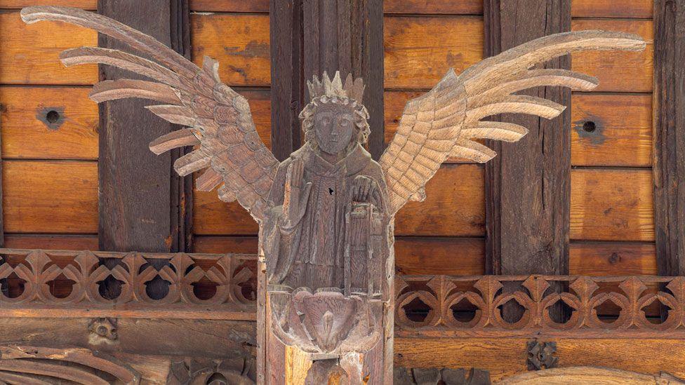 A wooden angel on the roof of St Wendreda's, March. It has a crown and its wings are extended in flight. Its right hand is raised in blessing and behind it can be seen the roof beams and planks