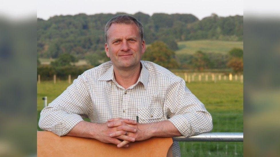 James Allen with a piece of their cow hide leather with his arms leaning on it and a metal fence. He has a checked shirt on and is standing in front of fields of grass with trees in the distance.
