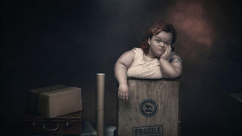 A woman with dwarfism has a bored expression on her face as she stands in a box marked fragile