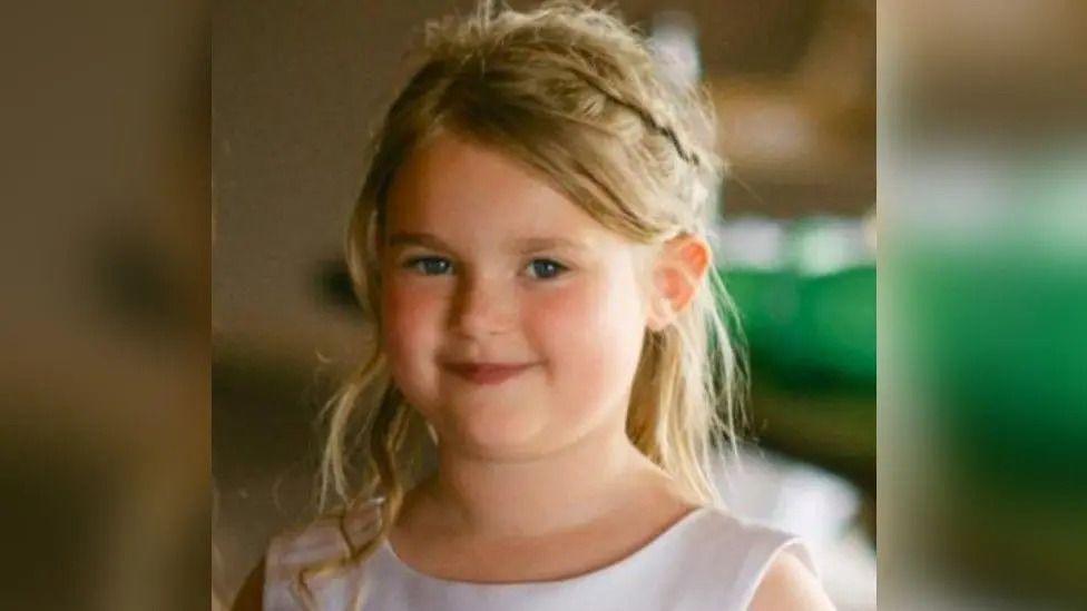 Elsie Gascoigne. She has blonde hair and blue eyes and is smiling at the camera. She wears a white dress.