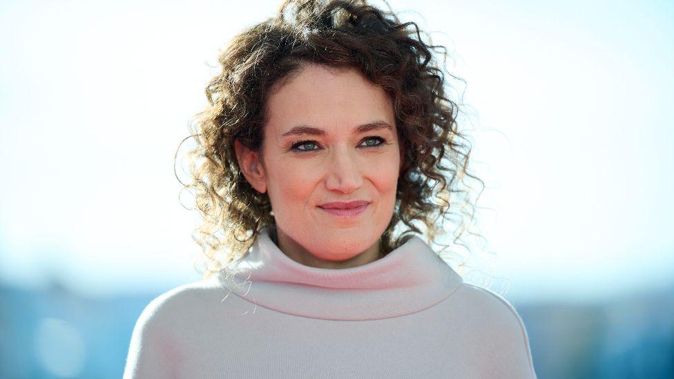 French director Coralie Fargeat attends a photocall of The Substance during Sitges Film Festival on 4 Ocboter, 2024, in Sitges, Spain. She has curly medium-length brown hair, blue eyes and is wearing a cream jumper.