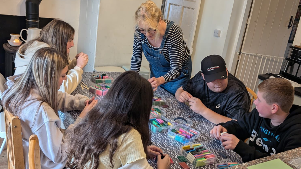 Three teenage girls and two teenage boys sat around a kitchen table. The girls have long brown hair, one of the boys has short fair hair and the other is wearing a black baseball cap. Boxes of coloured model clay are in the middle of the table and they are all chatting and moulding the clay. A blonde woman wearing a striped t shirt and denim dungarees stand next to them and leans over the table.