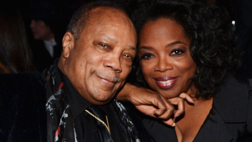 Quincy Jones and Oprah Winfrey sitting with their heads close together and smiling at the Rock and Roll Hall of Fame Induction Ceremony in 2013