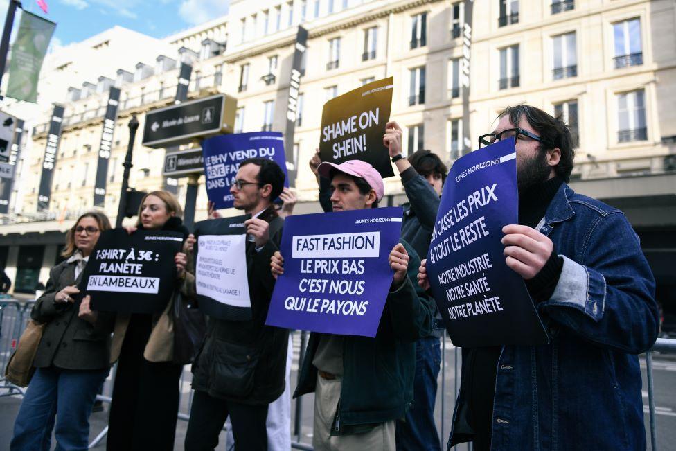 A group of protesters holding signs that say things like "fast fashion" and "shame on Shein" stand outside a row of buildings.