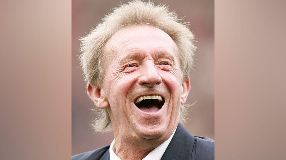 Denis Law with blond spiky hair laughing out loud and wearing a navy suit and white shirt