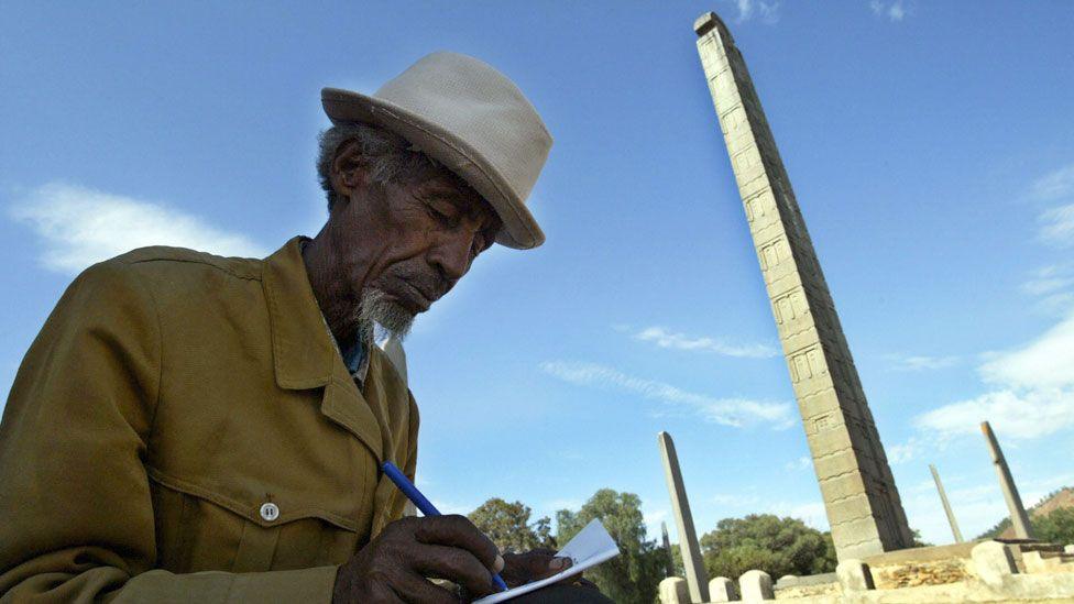 A man in a hat with grey hair and and goatee bear writes in a notebook, behind him are the obelisks of Aksum