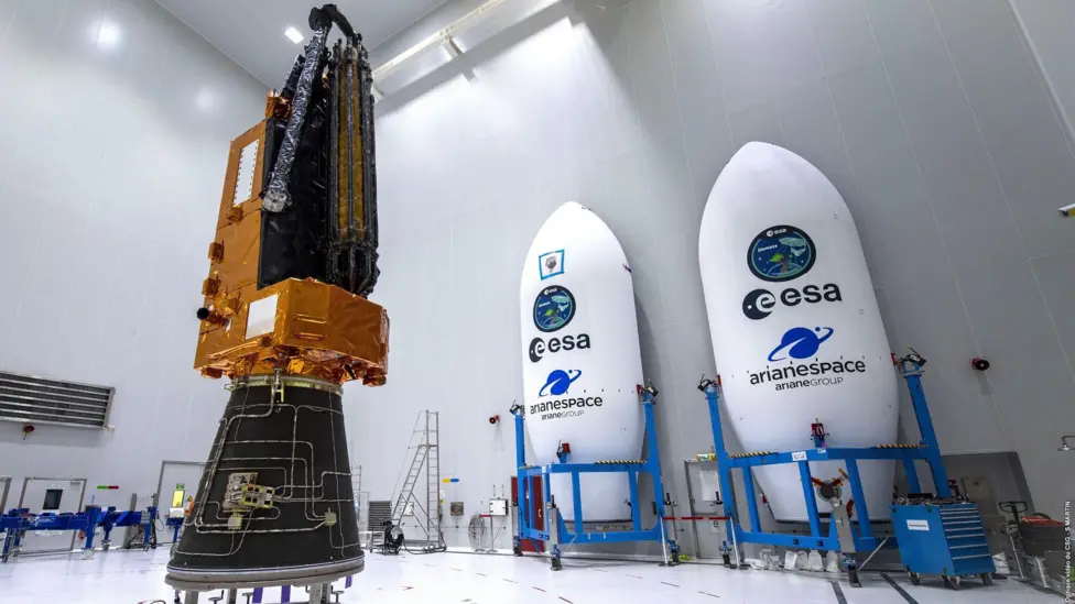 The biomass satellite sits inside an all-white warehouse. It has a black cone-like base, an orange l-shaped top that has the look of foil, with the antenna attached and furled. To the left of the satellite sit two shells which will hold the satellite, they are white elongated cones with ESA written on them, and held in blue scaffolding trolleys. 