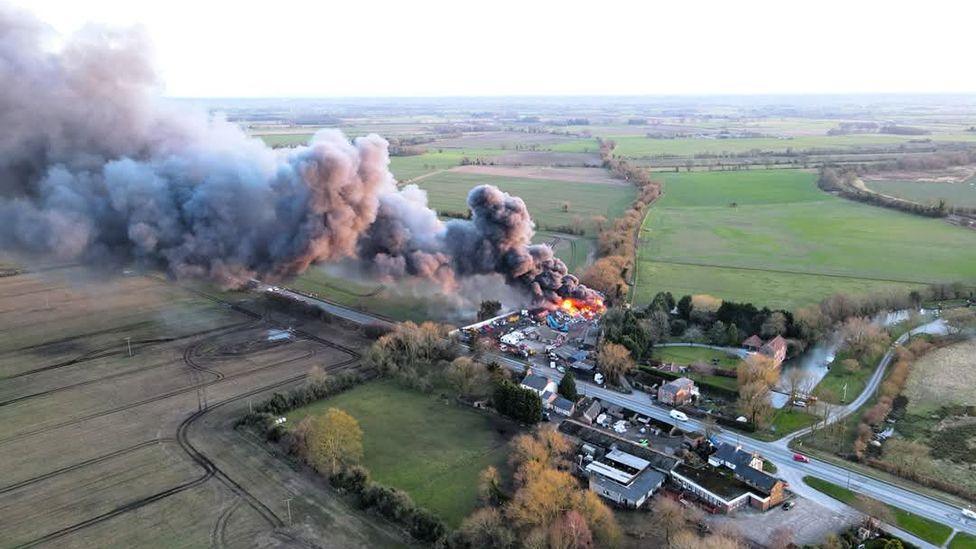 Aerial view of the fire showing flames and a large column of dark smoke drifting with buildings in the middle surrounded by green fields.