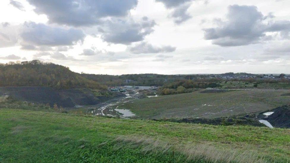 A wide view of the site showing undulating grassland and housing in the distance