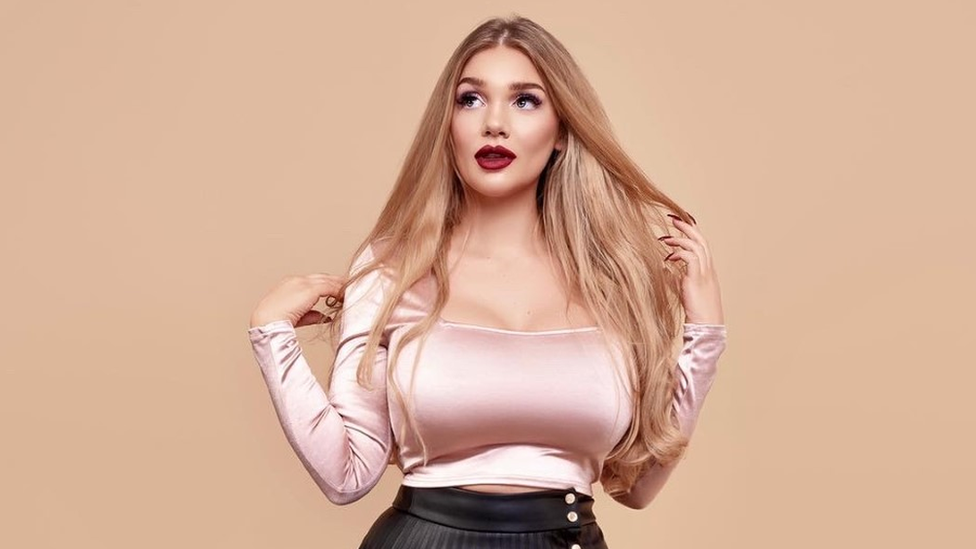 Lucy wears a pale pink silk top and has long blonde hair styled straight around her shoulders. She is wearing dark red lipstick.